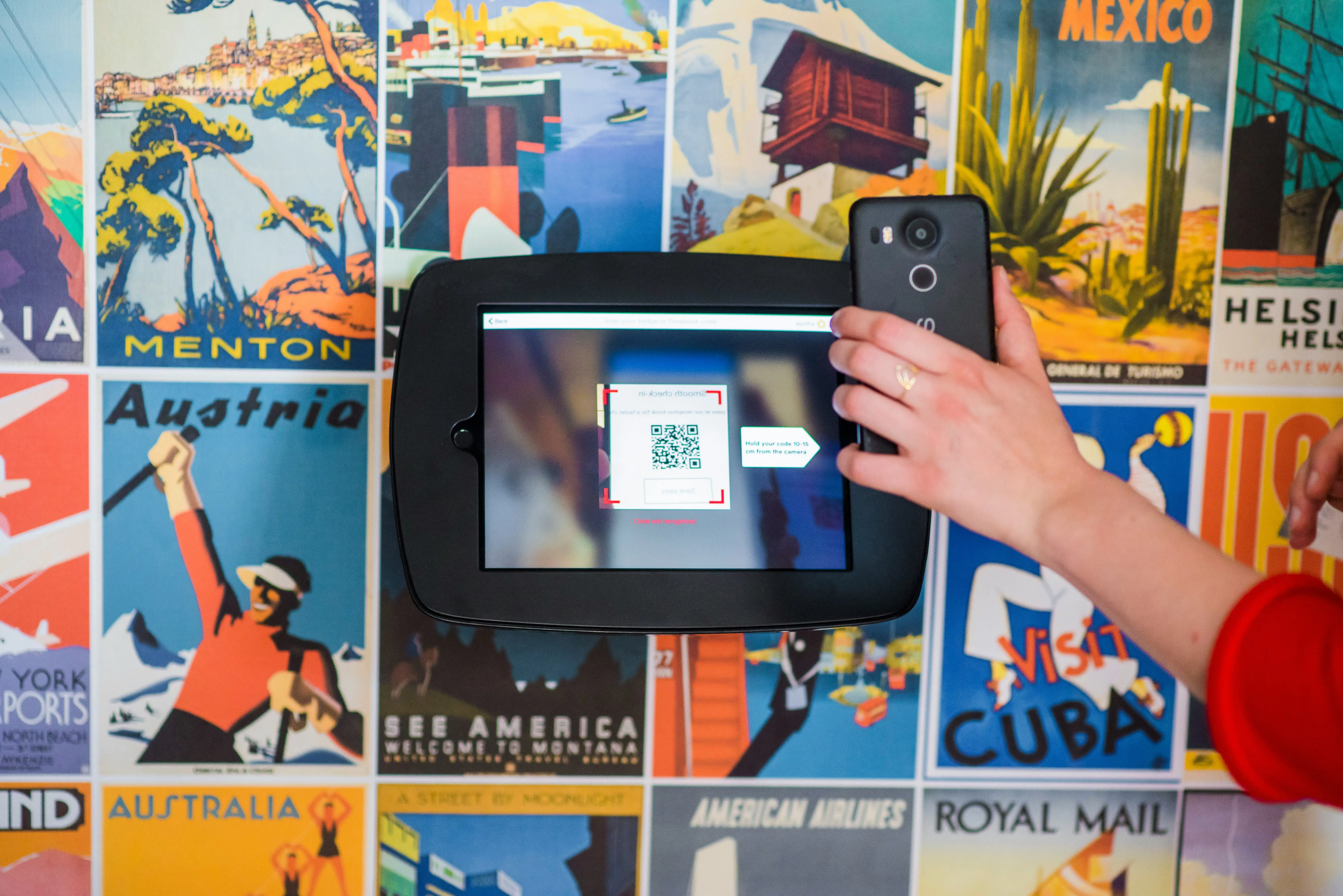 A person uses a smartphone to scan a QR code displayed on a tablet screen, with a colorful wall of vintage travel posters in the background.