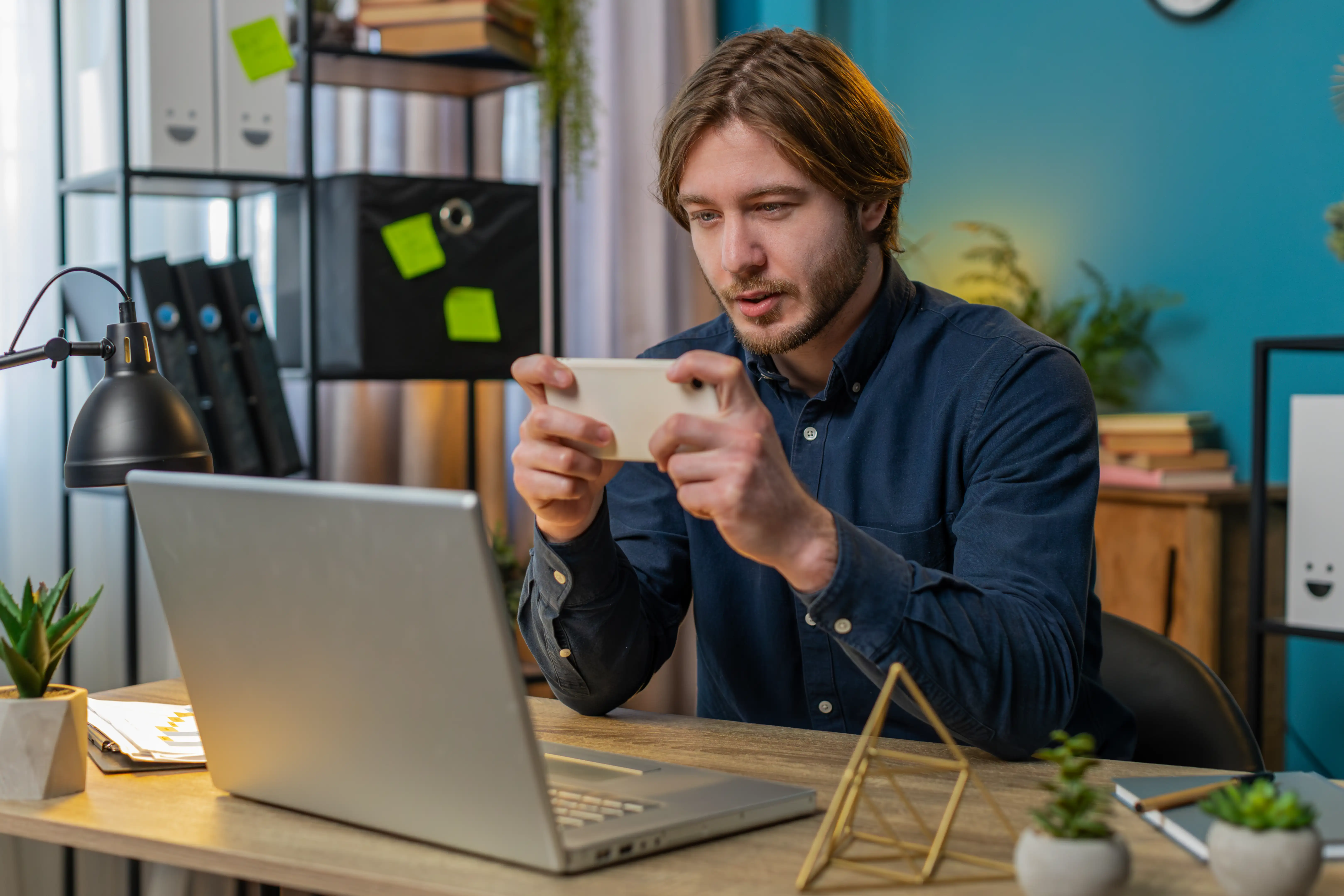Professional man in blue shirt scanning QR code with smartphone while working at laptop in modern home office with organized shelves and plants for remote work and mobile technology