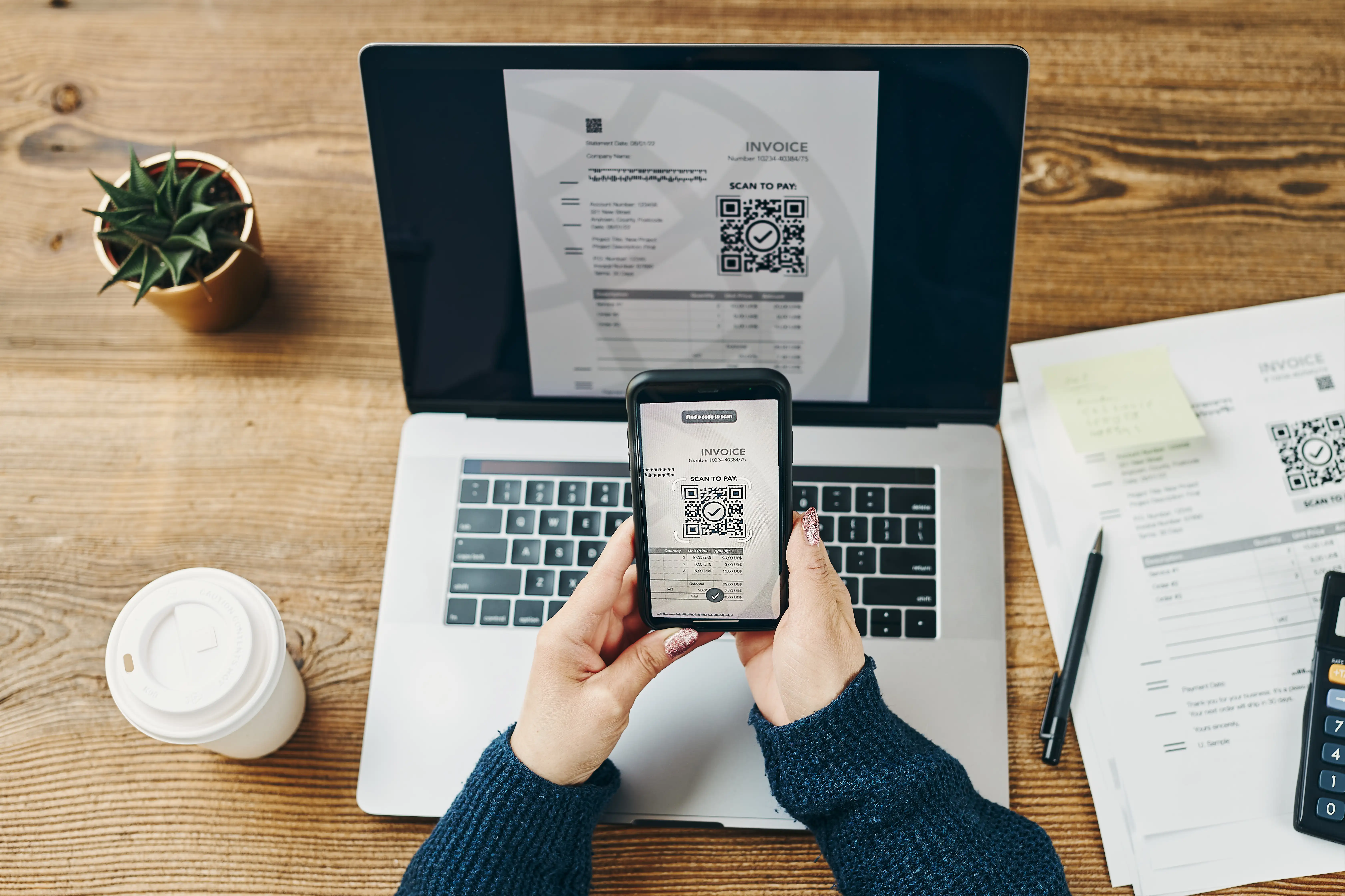 Overhead view of hands holding smartphone scanning QR code on digital invoice displayed on laptop screen with coffee and paperwork on wooden desk for mobile payment processing