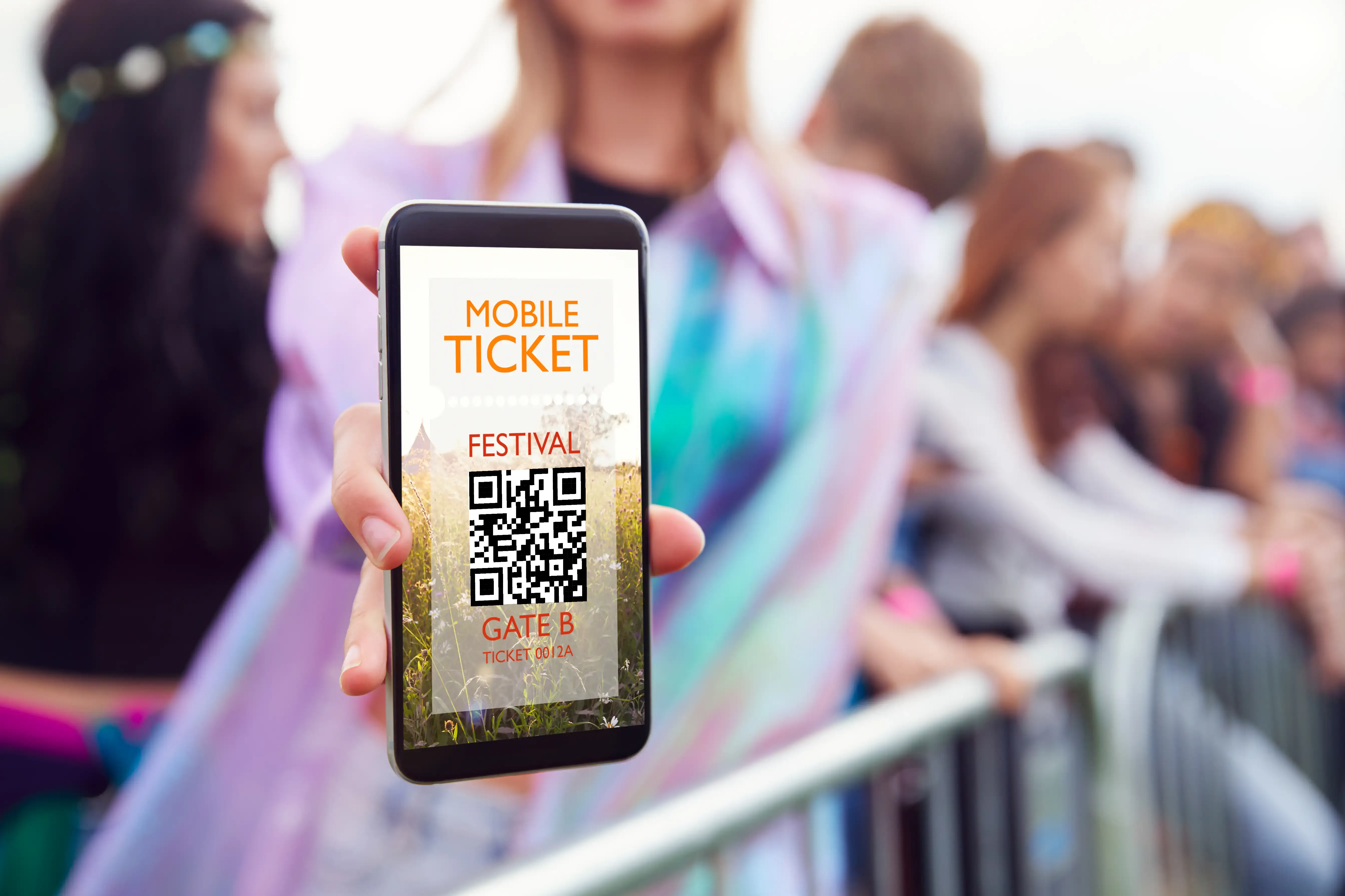 Hand holding smartphone displaying mobile festival ticket with QR code for Gate B entry at outdoor music festival with colorful blurred crowd in background for digital ticketing and event access