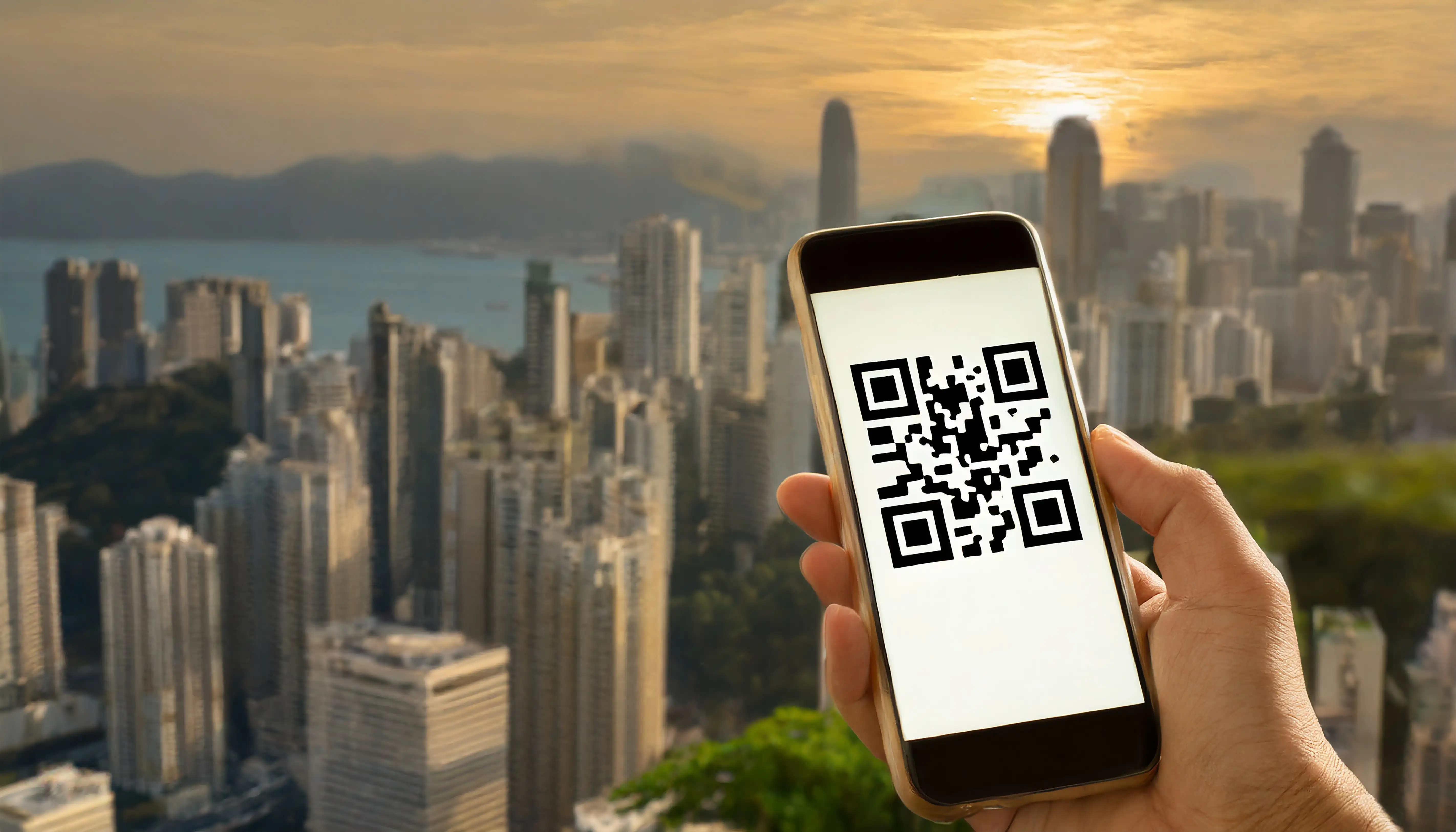 Hand holding smartphone displaying QR code with Hong Kong urban skyline and harbor at golden sunset for tourism, travel guide or location-based mobile app services