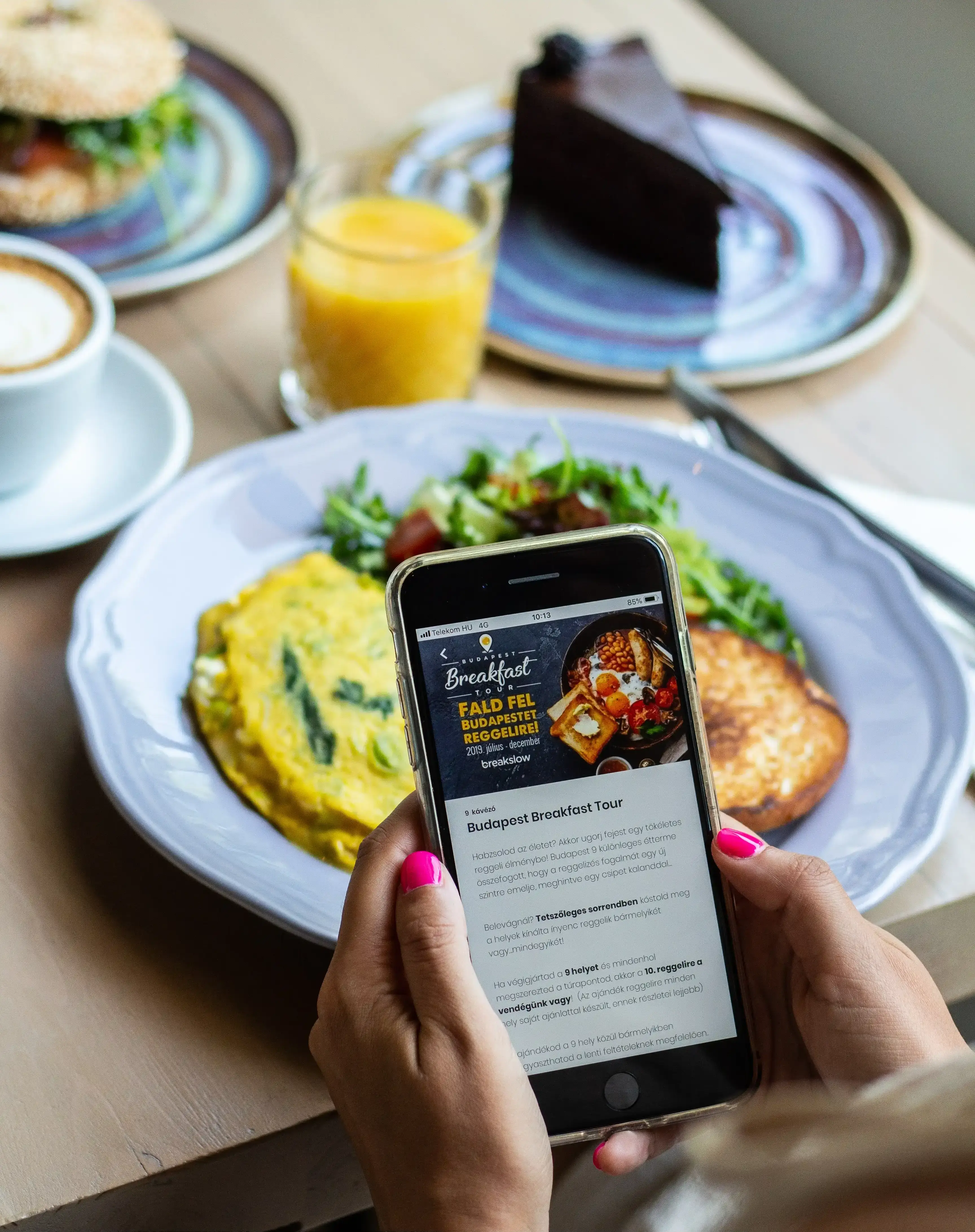 Woman with pink nails holding smartphone displaying Budapest Breakfast Tour booking app over restaurant table with omelet, salad, coffee, orange juice and chocolate cake for food tour reservation and dining experience