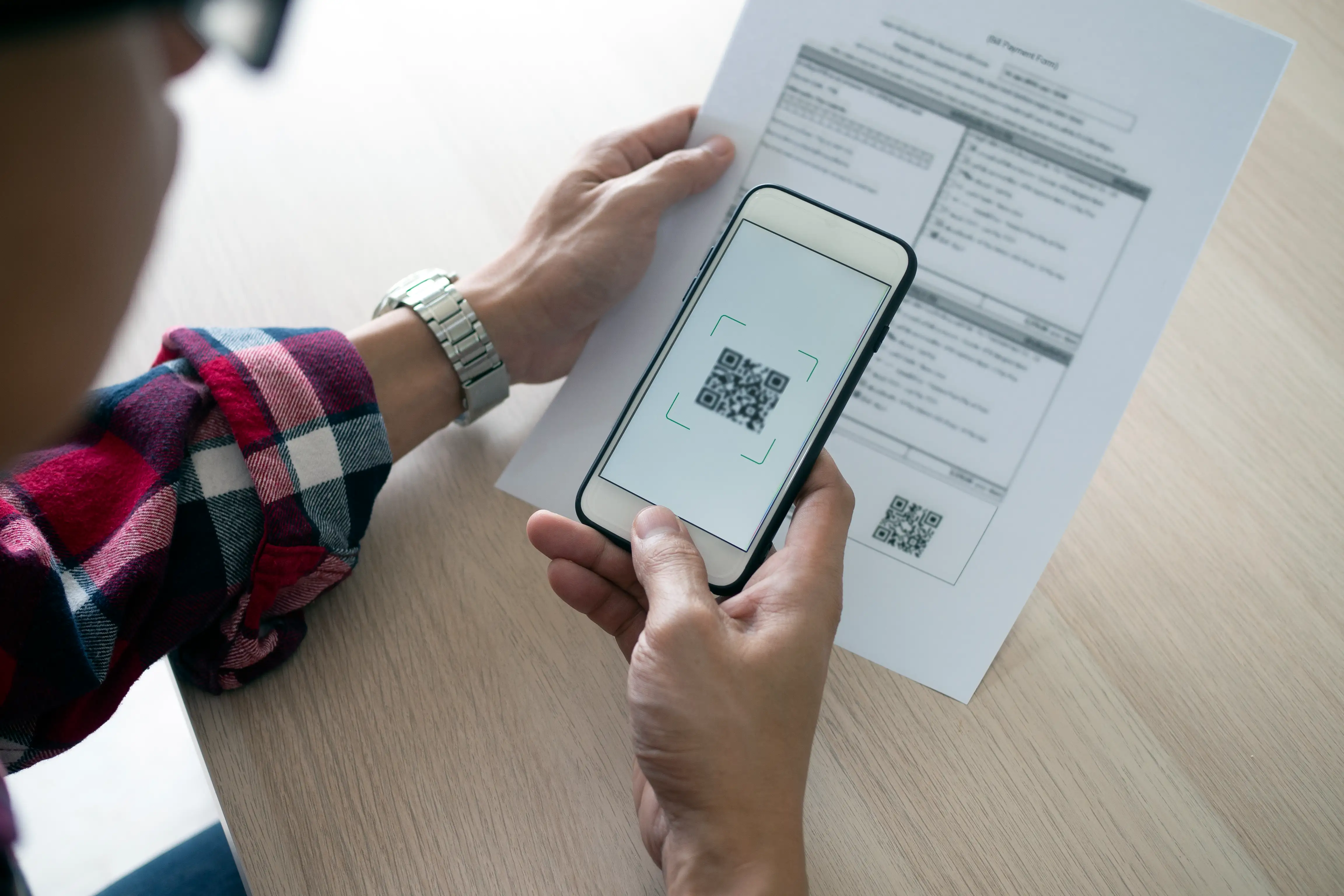 Overhead view of person in plaid shirt scanning QR code on printed document with smartphone for paperless verification, digital document access and mobile scanning application