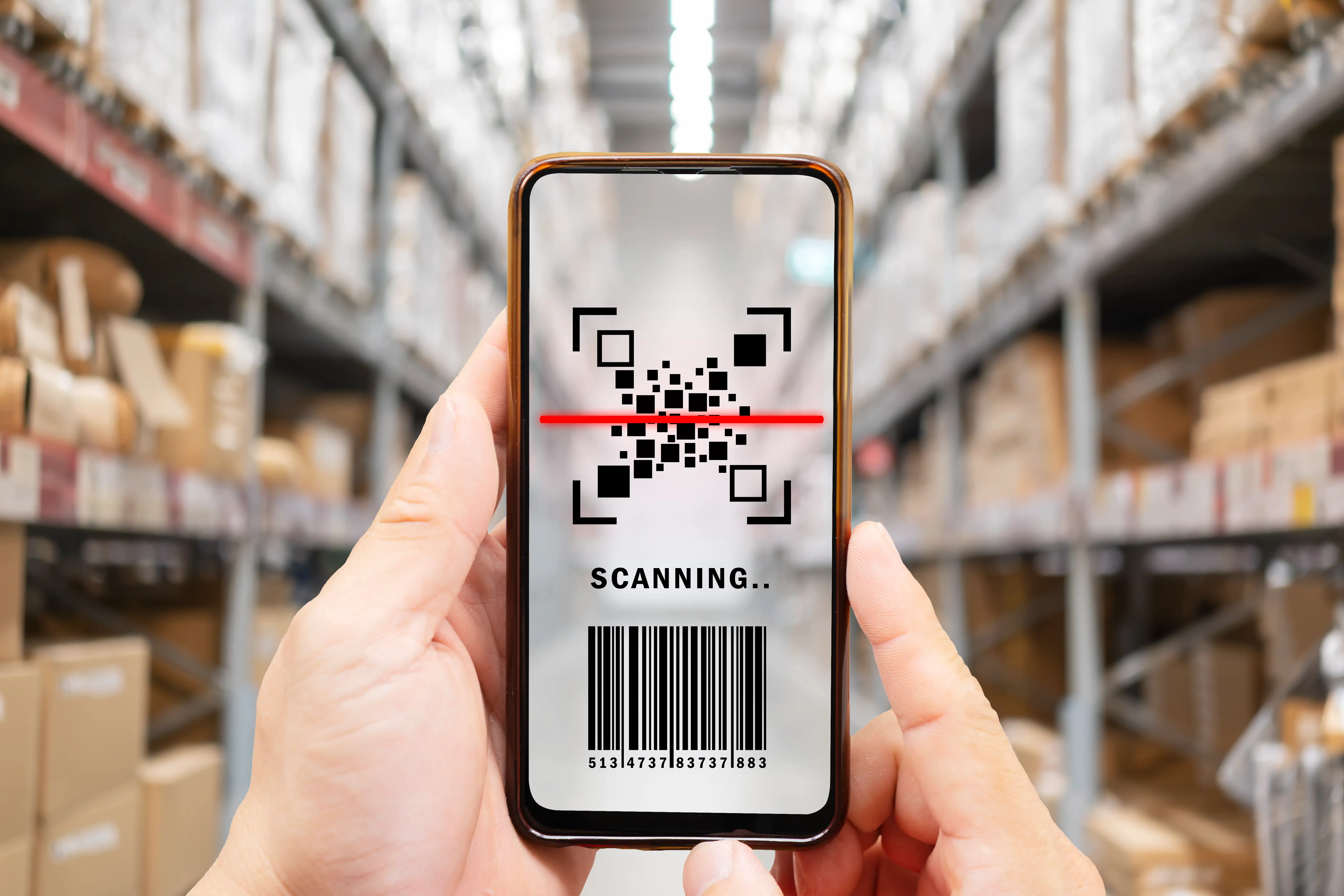 Hand holding smartphone scanning barcode and QR code with red laser line in warehouse with shelving units for inventory management, logistics tracking and supply chain operations
