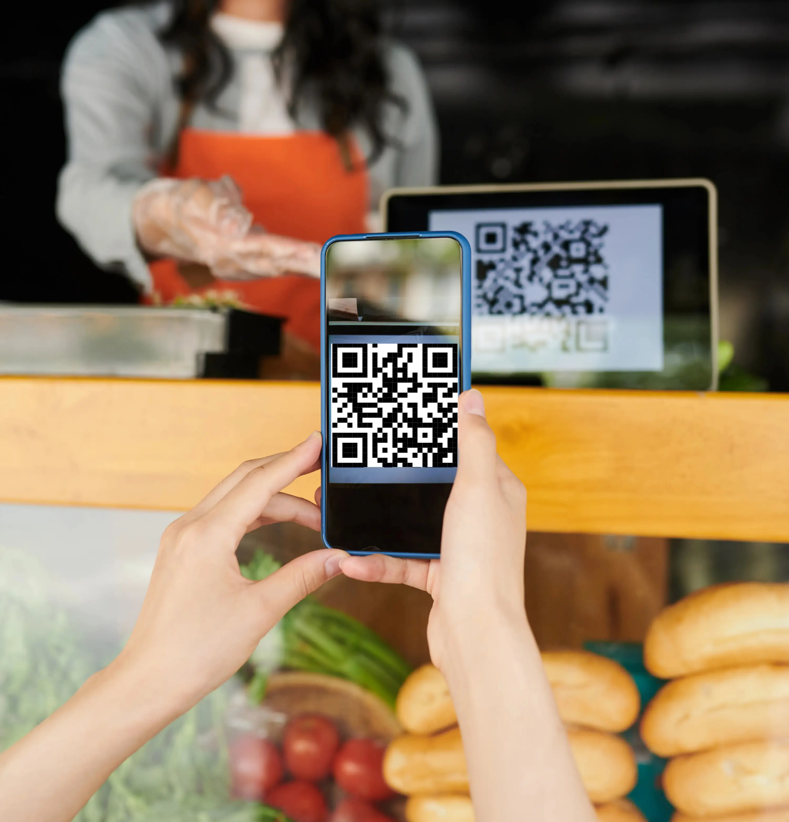 Customer scanning QR code with smartphone at grocery store checkout counter with cashier in orange apron, fresh vegetables and bread display for contactless payment and retail transaction