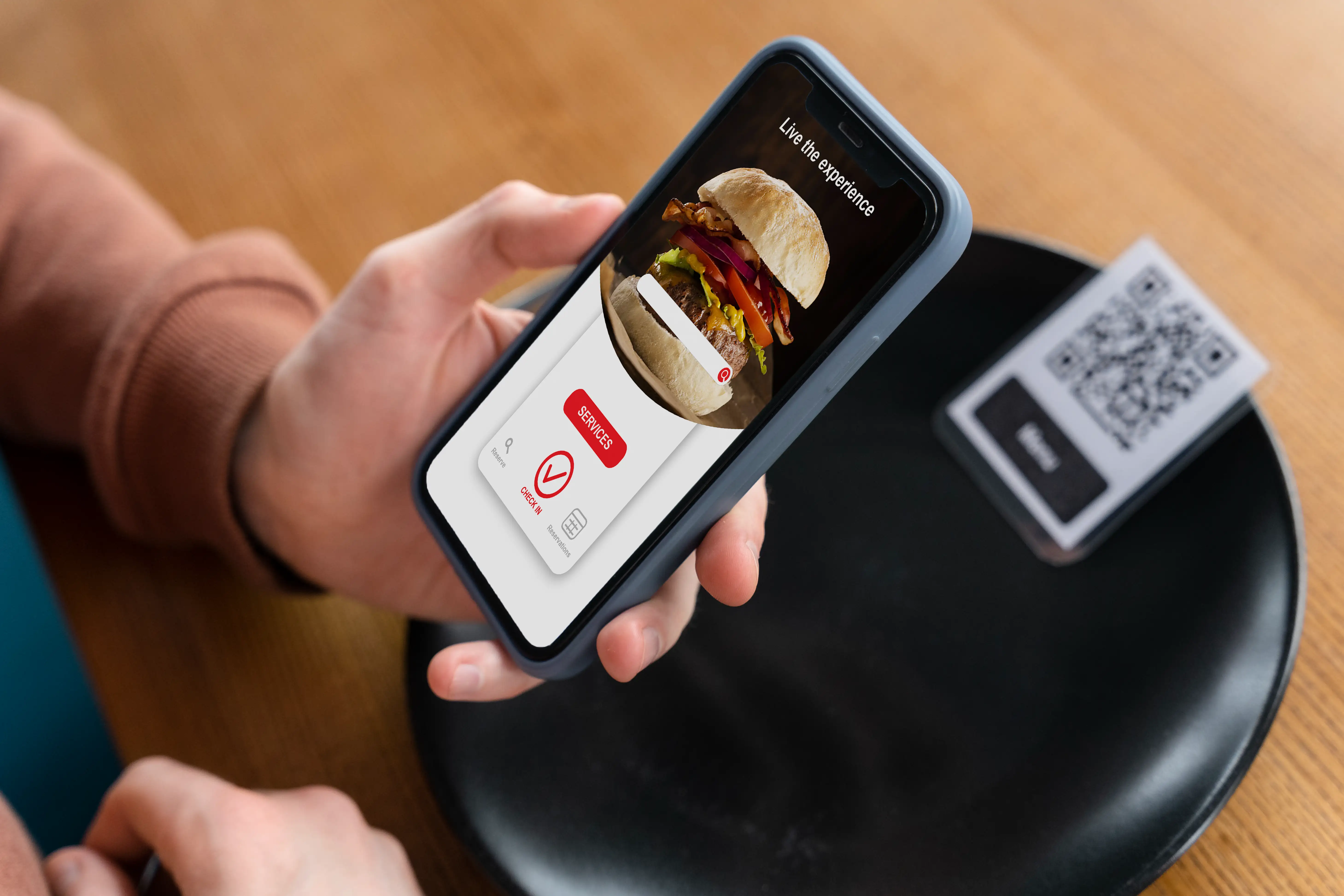 Hand holding smartphone showing burger food delivery app with red services button and QR code scanner on table for contactless restaurant ordering and live dining experience