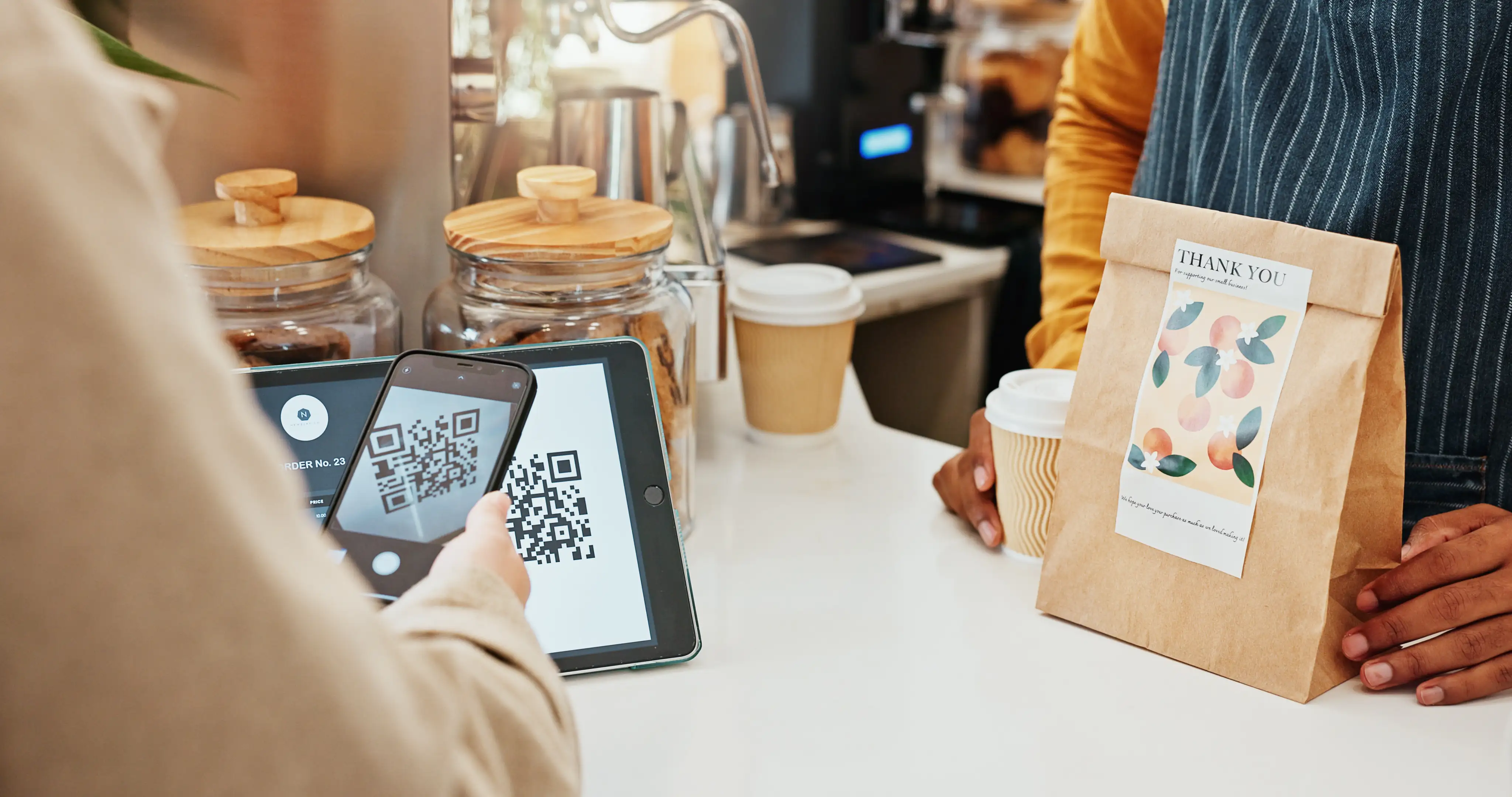 Customer scanning QR code payment at cafe counter with tablet while barista hands over takeout coffee and thank you bag for contactless transaction at coffee shop