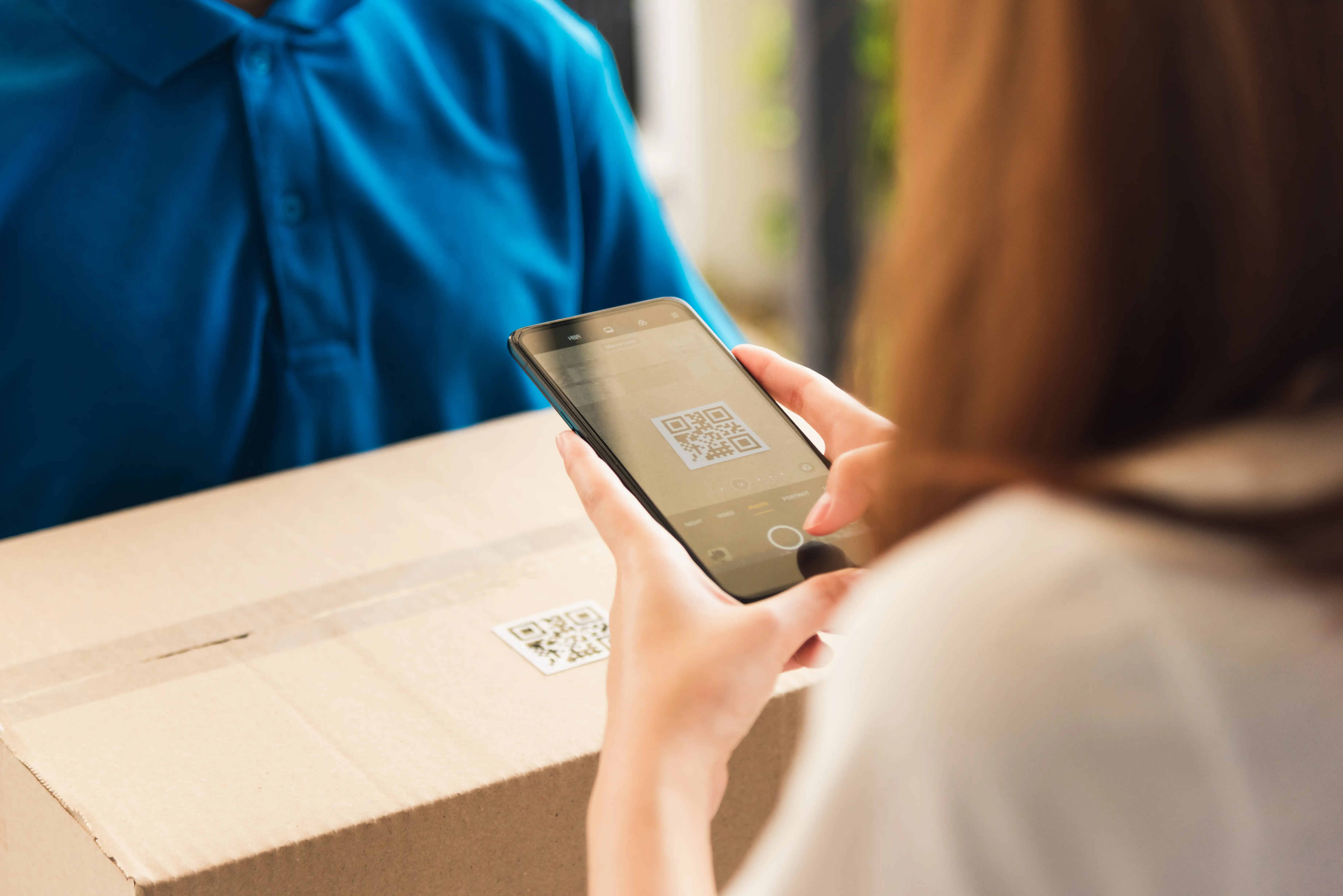 Person in blue shirt scanning QR code on cardboard shipping box with smartphone for package tracking and delivery verification in home setting