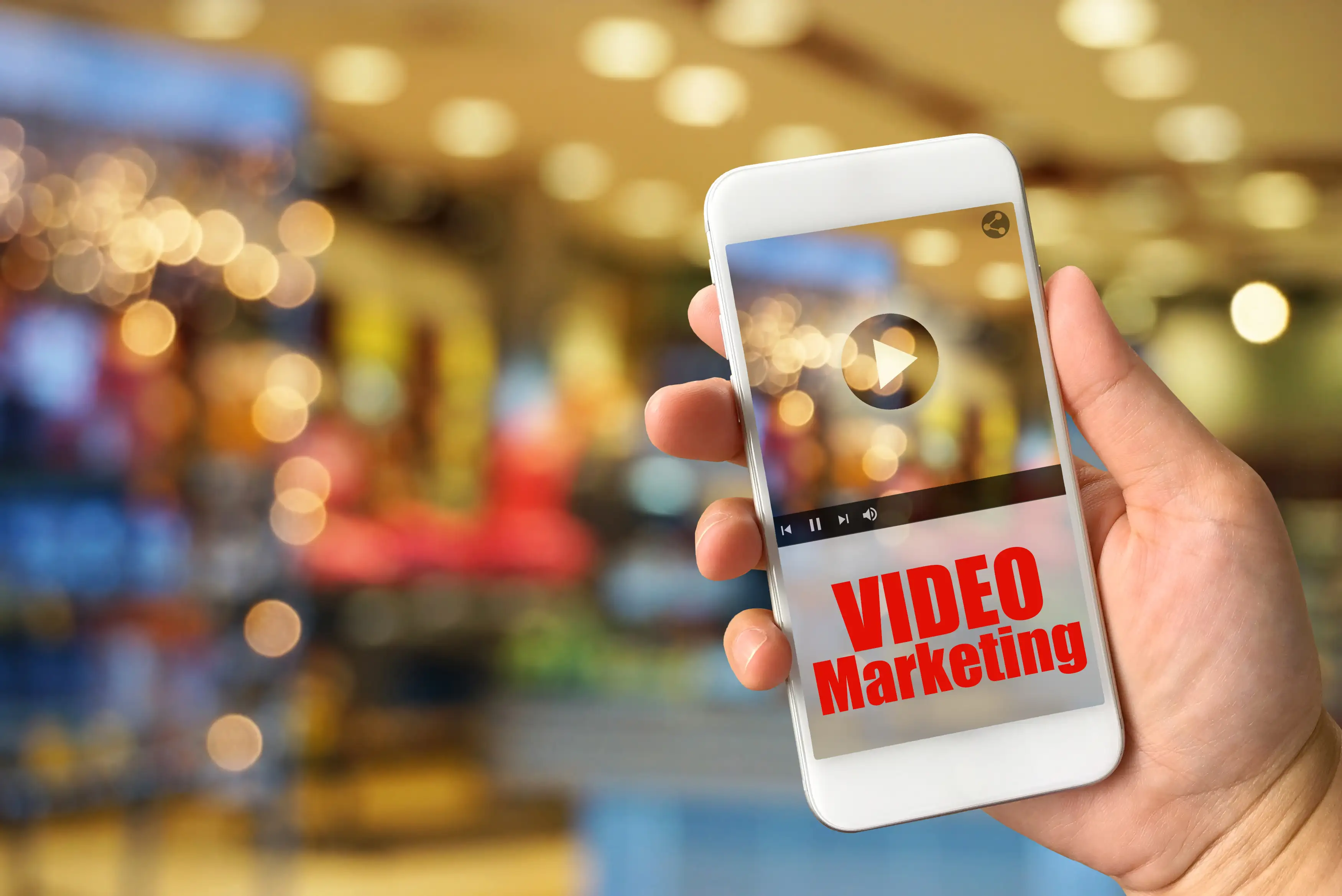 Hand holding white smartphone displaying video marketing content with play button and bokeh shopping mall background for mobile video advertising and digital marketing campaigns