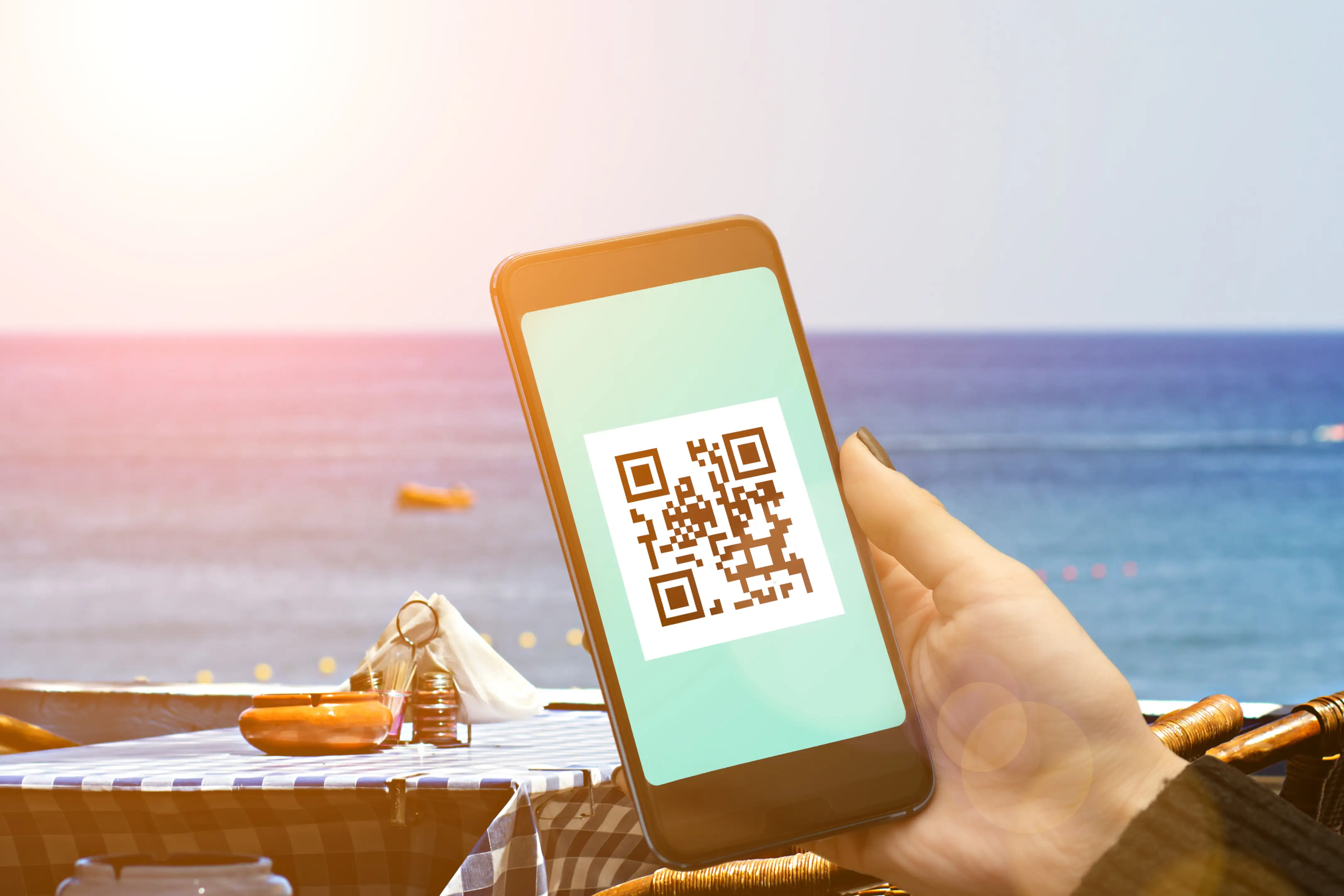 Alt Description: Hand holding smartphone scanning brown QR code at ocean beach location, demonstrating travel and tourism QR code usage for vacation activities and information